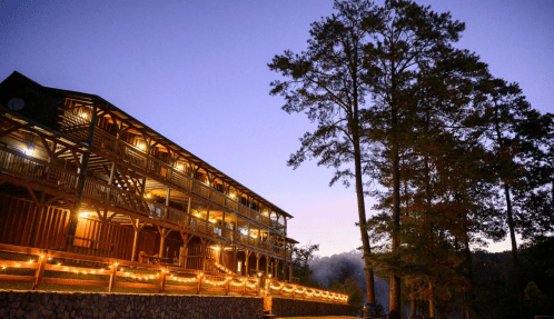 A rustic lodge illuminated by warm lights at dusk, surrounded by tall trees and a serene atmosphere.