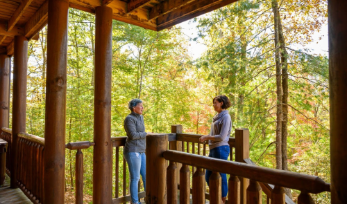 Two women converse on a wooden porch surrounded by vibrant autumn trees.