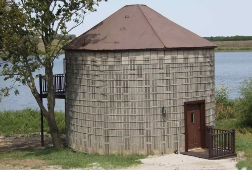 A round, stone building with a brown roof, near a river, surrounded by greenery and a tree.