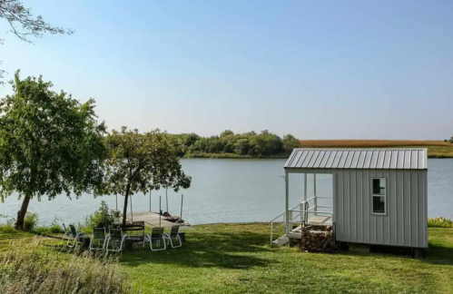 A serene lakeside scene featuring a small gray cabin, chairs, and trees under a clear blue sky.
