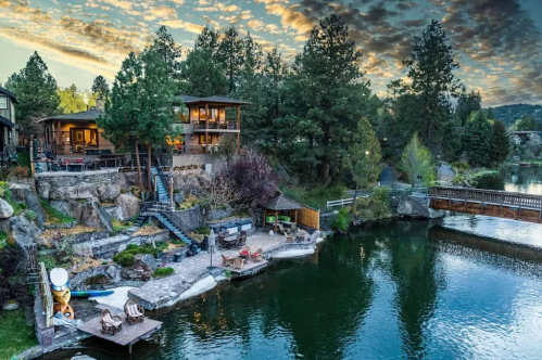 Scenic lakeside view with a wooden deck, lush trees, and a cozy cabin under a colorful sky at sunset.