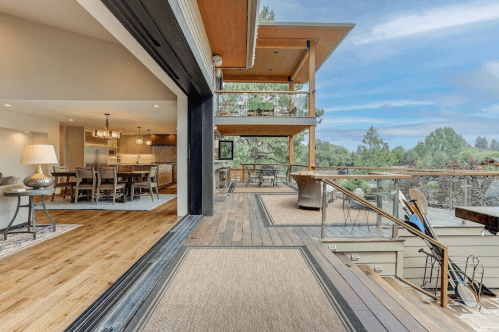 A modern home interior opens to a spacious deck with scenic views, featuring wooden floors and stylish furnishings.