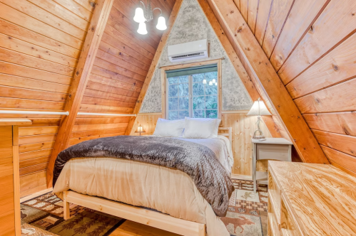 Cozy A-frame bedroom with wooden walls, a plush bed, and warm lighting, featuring a window with a forest view.