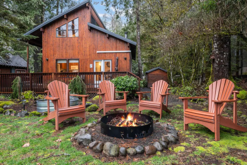 A cozy wooden cabin surrounded by trees, featuring a fire pit and four Adirondack chairs in a serene outdoor setting.