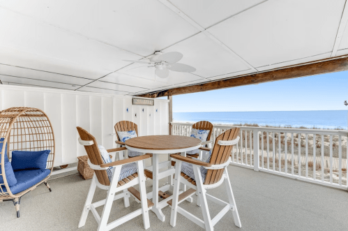 Cozy beachfront patio with a round table, chairs, and a hanging chair, overlooking the ocean and sandy shore.