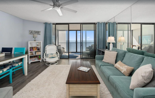 Bright living room with teal furniture, a coffee table, and large windows showing a view of the ocean. Cozy and inviting space.