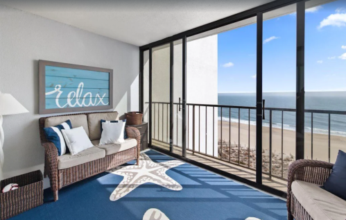 Cozy beachside balcony with a sofa, starfish rug, and ocean view through large glass doors.