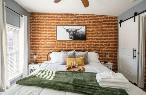 Cozy bedroom with a brick wall, a large bed, decorative pillows, and a cow print above the headboard.