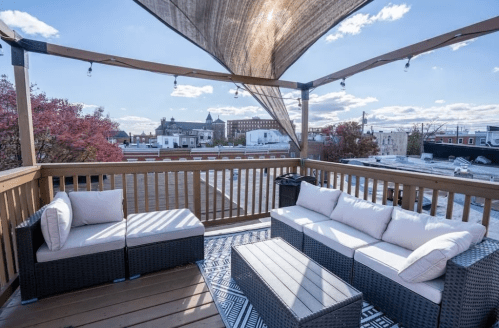 A rooftop patio with a shaded seating area, featuring a sectional sofa and coffee table, overlooking city buildings.