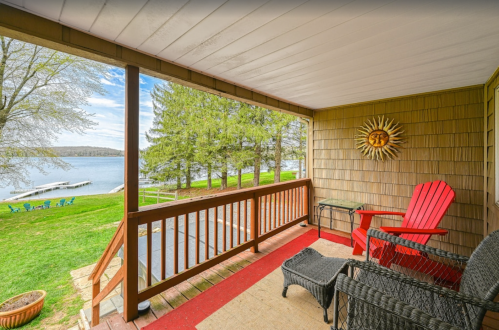 A cozy porch with red chairs overlooks a serene lake and green trees on a sunny day.