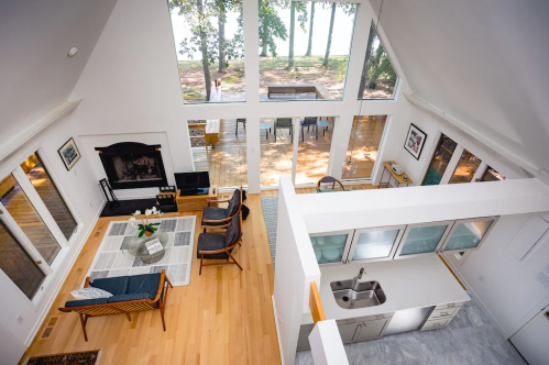 Bright, modern living space with large windows, wooden floors, and a view of trees outside. Cozy seating area and kitchen.