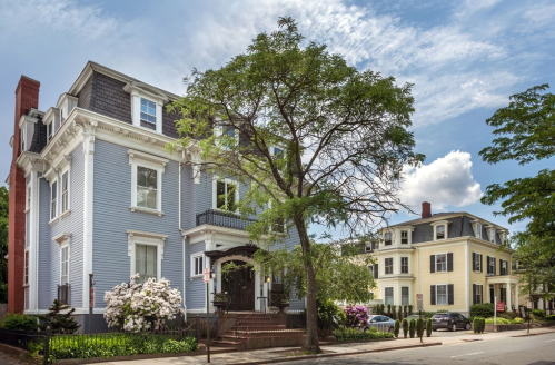 A charming blue house with white trim and a tree in front, alongside another elegant home under a clear blue sky.