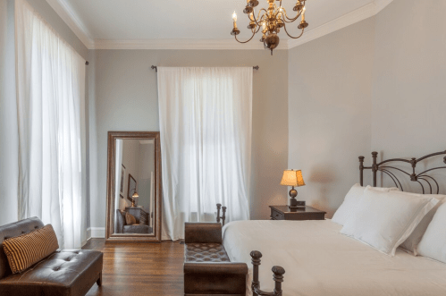 Bright bedroom with a bed, mirror, armchair, and chandelier, featuring light curtains and wooden flooring.