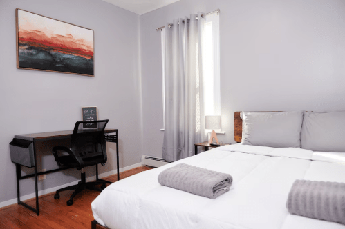 A cozy bedroom with a bed, desk, chair, and large window, featuring soft gray tones and a colorful wall art piece.