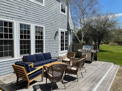 A patio with a blue sofa, wooden table, wicker chairs, and a grill, surrounded by greenery and a clear sky.