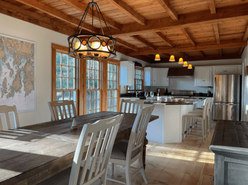 Bright kitchen with wooden beams, a large dining table, and a bar area, featuring a map on the wall and natural light.
