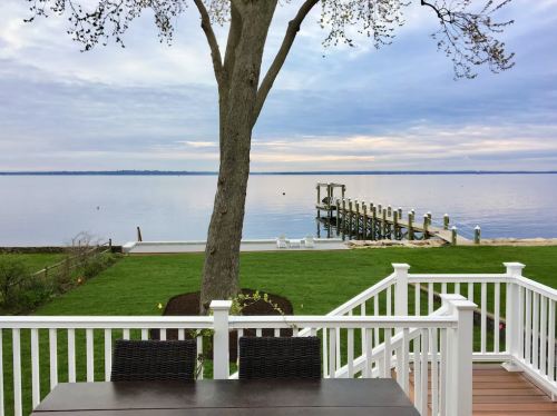 A serene lakeside view from a deck, featuring a tree, dock, and calm water under a cloudy sky.