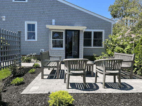 A cozy outdoor seating area with wooden chairs and a stone patio, surrounded by greenery and a house in the background.