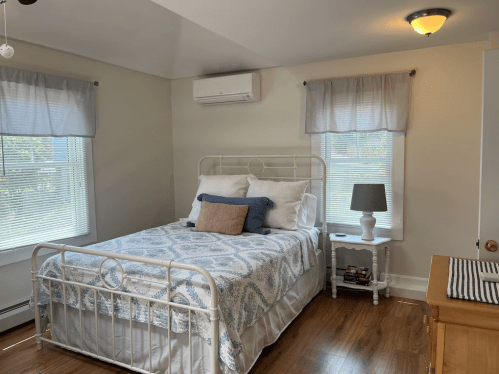 Cozy bedroom with a white metal bed, light blue bedding, two windows, and a small nightstand with a lamp.