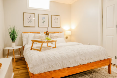 Cozy bedroom with a wooden bed, white bedding, decorative wall art, and a tray with wine and grapes on a bedside table.