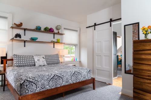 Cozy bedroom featuring a patterned bedspread, wooden shelves, and a sliding door, with natural light streaming in.