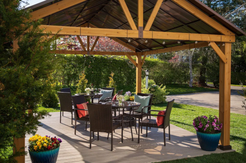 A cozy outdoor patio with a wooden gazebo, dining table, chairs, and colorful flower pots.