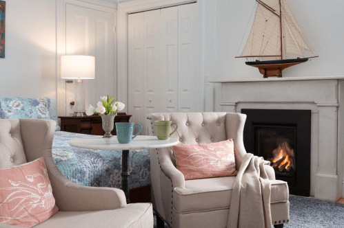 Cozy room with two armchairs, a small table, a bed, and a fireplace, featuring a decorative sailboat and flowers.