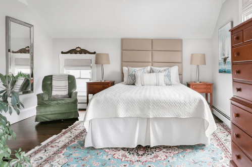 Cozy bedroom featuring a large bed, green armchair, wooden dressers, and a colorful area rug. Bright and inviting decor.