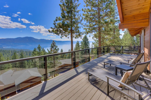 A scenic deck with lounge chairs overlooking a lake and mountains, surrounded by tall pine trees under a clear blue sky.