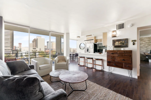 Modern living room with a cozy sofa, coffee tables, and a kitchen area, featuring large windows and city views.