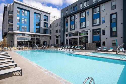 Modern apartment complex with a large outdoor pool and lounge chairs, surrounded by stylish buildings and clear blue skies.