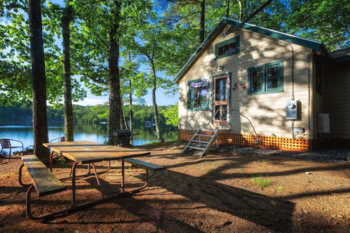 A cozy lakeside cabin surrounded by trees, with a picnic table in front and a serene lake in the background.