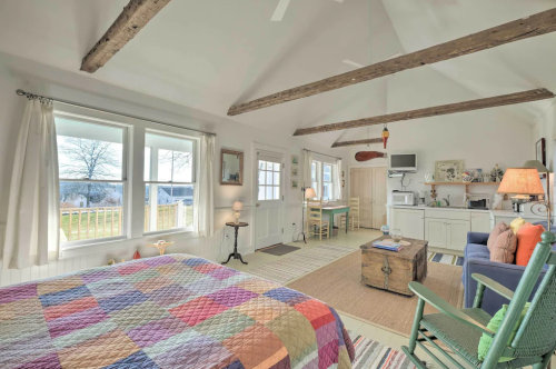 Cozy bedroom with a colorful quilt, wooden beams, large windows, and a sitting area with a green rocking chair.