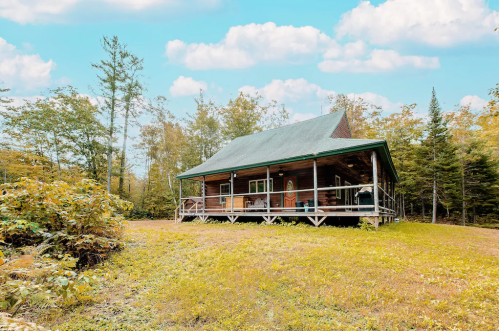 A cozy wooden cabin with a green roof, surrounded by trees and a grassy area under a blue sky with fluffy clouds.