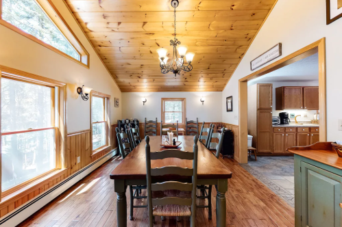 A cozy dining area with a wooden table, chairs, and a chandelier, featuring large windows and a view of the kitchen.