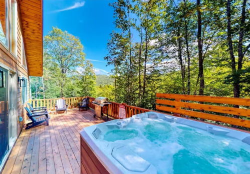 A wooden deck with a hot tub, grill, and seating, surrounded by trees and a clear blue sky.