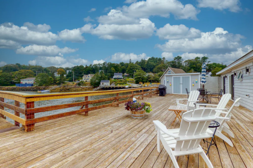 A wooden deck with white chairs overlooks a calm waterfront, surrounded by trees and houses under a blue sky with clouds.