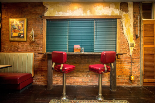 Two red bar stools at a small counter in a rustic restaurant with exposed brick walls and a window with blinds.