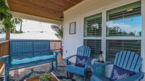 A cozy porch with blue seating, a patterned rug, and decorative pillows, surrounded by greenery and natural light.