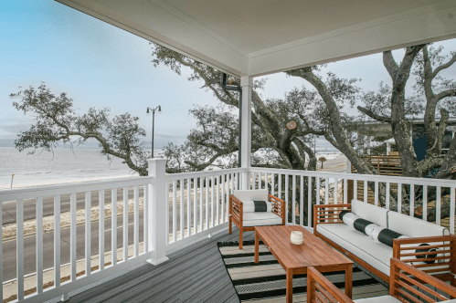 Cozy porch with wooden furniture overlooking a beach and trees, creating a serene coastal atmosphere.
