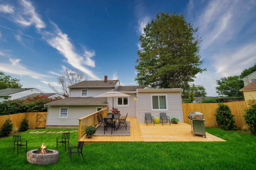 A backyard with a wooden deck, seating area, grill, and fire pit, surrounded by green grass and a blue sky.