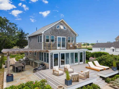A modern beach house with a spacious deck, outdoor seating, and lush greenery under a clear blue sky.