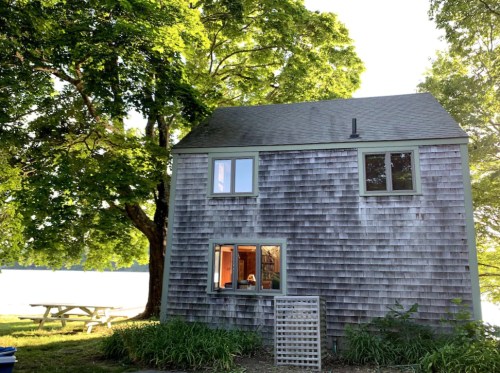 A rustic two-story house with wooden siding, surrounded by trees, near a lake and picnic area.