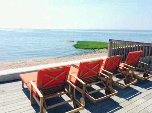 Four orange lounge chairs lined up by a calm waterfront, with green grass and a clear blue sky in the background.