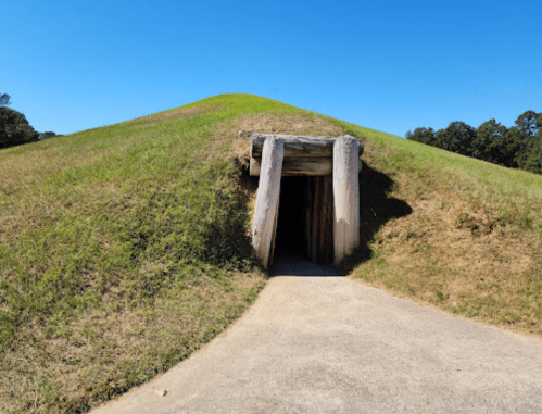 A grassy mound with a wooden entrance, leading into a dark tunnel, under a clear blue sky.