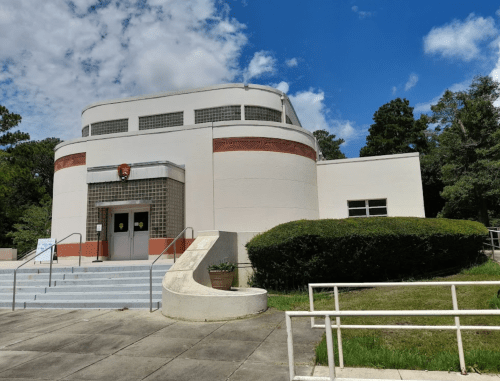 A modern, round building with large windows, surrounded by greenery and a clear blue sky.