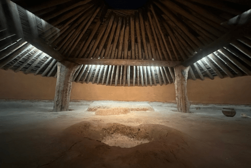 Interior of a traditional round structure with a wooden roof, featuring a central pit and soft lighting.
