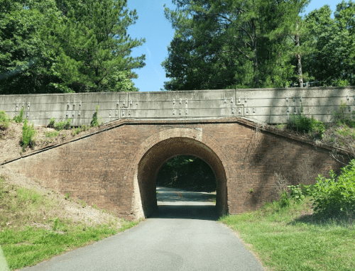 A brick arch bridge with a concrete wall above, surrounded by greenery and a clear blue sky.