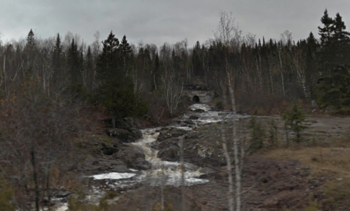A rocky stream flows through a forested area with sparse trees and a cloudy sky in the background.