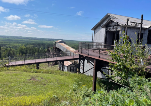 Abandoned industrial building on a hillside, surrounded by greenery and under a clear blue sky.
